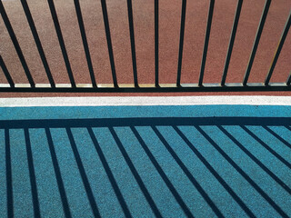 Black metal fence and its shadow on a colored covering of a sport playground