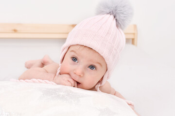 Thoghtful, adorable happy caucasian four-five months baby in winter knitted hat lying on bed. Baby care and winter clothes concept.