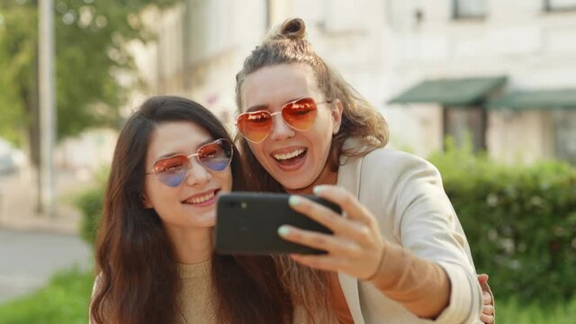 Positive emotions and love. Two young smiling hipster blond women taking selfie self portrait photos on smartphone. Pretty stylish girls on street background. LGBTQI, Pride Event, LGBT Pride Month