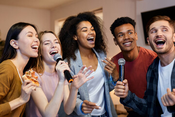 Sing it. Enthusiastic group of friends singing with microphone while playing karaoke at home,...