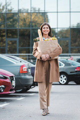  woman in autumn outfit walking along car parking while carrying shopping bag with food