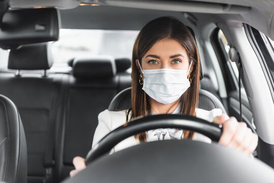 woman in mask looking at camera while driving car on blurred foreground