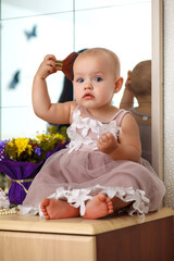 Fototapeta premium A cute little girl is holding a makeup brush and having fun at home. Kid's fashion. Little girl fashionista. Nice cozy bedroom. Beautiful girl sitting on a table with makeup tools.