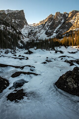 A view of Dream Lake in Rocky Mountain National Park with snow covered peaks surrounded by a forest of trees, snow, and ice. Shot is taken with amongst rocks and a frozen stream.