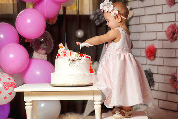 Cute smiling baby girl in pink dress with her first birthday cake. One year old baby celebrates birthday. Cute dress in pink color. Happy birthday card.