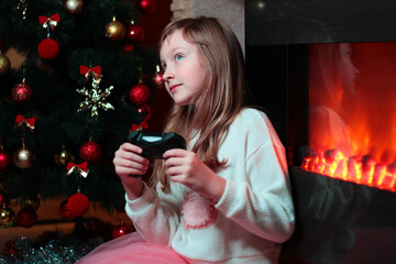A girl plays a game console on the background of a Christmas tree and a fireplace. Gamer. New year's entertainment.