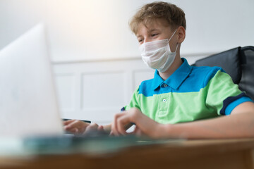 Fototapeta na wymiar Handsome European boy in a disposable face mask is doing homework on his laptop during quarantine. Online studying concept