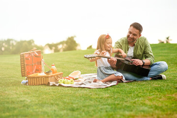 Full length shot of happy little girl sitting with her loving father on a green grass in park and learning how to play guitar