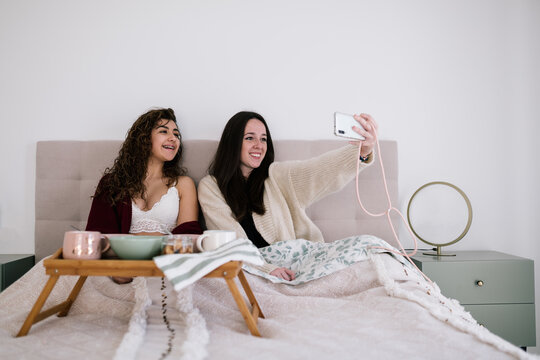 portrait of two white teenage girls in lingerie and cardigan on the bed taking a selfie with their mobile phone. Above the bed is a small breakfast table with cups and a bowl