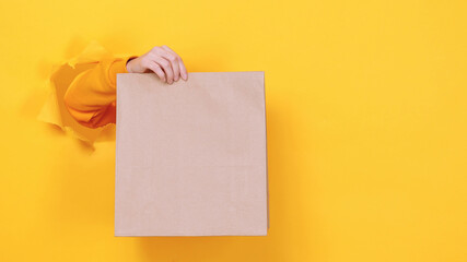 Close up womale hold in hand brown clear empty blank craft paper takeaway bag isolated through torn yellow background. Packaging template mockup. Delivery service concept. Copy space Advertising area.