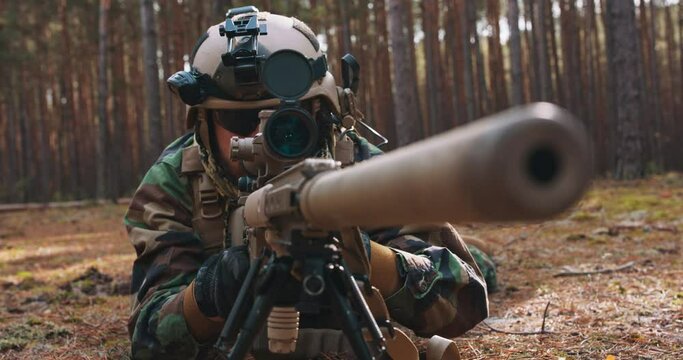 A bearded soldier lies on the forest litter in a tactical military uniform, with a helmet on his head, with a sniper rifle and opens the reticle and aims at the enemy through the scope