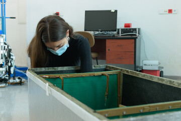 A young female engineer with dark hair and wearing a mask and a black sweater fixing an...