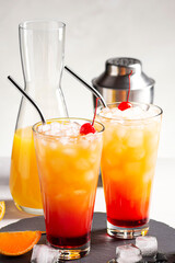 Two alcoholic cocktails tequila Sunrise with ice on a gray concrete background, next to a shaker and a decanter with juice. Close-up, vertical orientation, selective focus.