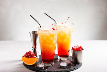 Ice cocktails tequila sunrise on a gray concrete table, next to a jigger and a cocktail cherry, horizontal orientation, selective focus, copy space.