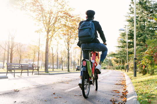 rear shot photograph of a student boy with a bag on his back carrying a red shared electric bike along a tree-lined park path at sunset