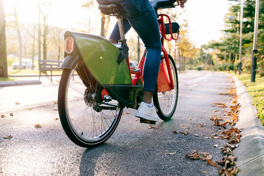 rear shot photograph of a student boy with a bag on his back carrying a red shared electric bike along a tree-lined park path at sunset