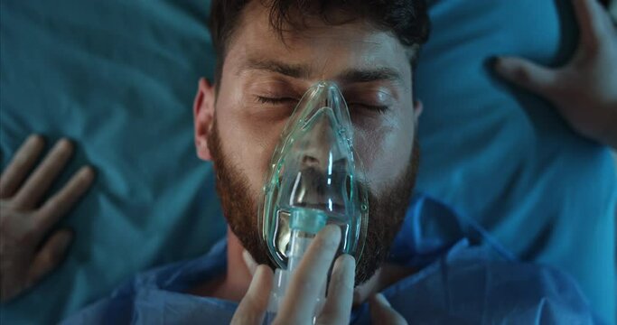 Close up of paramedics take off ill bearded guy face oxygen inhalation mask. Man in 40s losing consciousness while lying on medical transportation stretcher while going to hospital