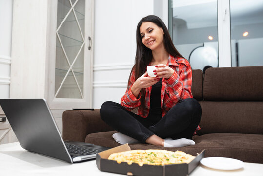 smiling girl with a cup of drink looks at the laptop while sitting on the couch