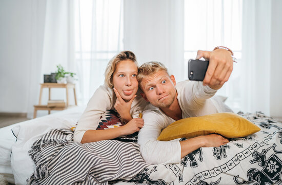 Cheerful young adults сouple in pajamas taking a Funny grimace selfie photo using a modern smartphone as they lazy relaxing lying in a cozy bed in the bedroom. . Couples relations concept image.
