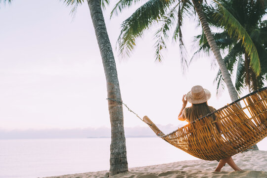 A young woman in straw hat sitting in a hammock swinging between a palm trees on the overseas island sand beach at sunrise time. Koh Samui, Thailand. Exotic vacation concept image.