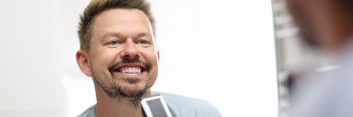 Smiling man stands in front of a mirror and holds shaving razor in his hands. How and when to shave...