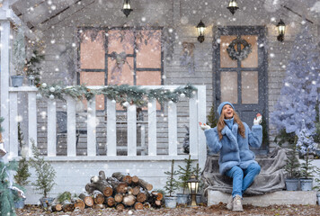 Happy young girl model is sitting on the porch of the Christmas house and catches the snow. Happy...