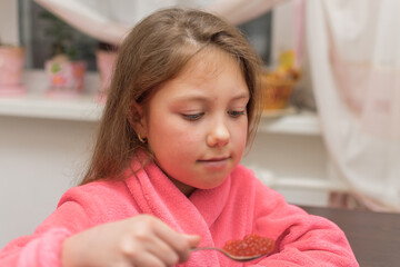 Fototapeta premium Children's portrait in the kitchen. A cute little girl eats red caviar with a spoon. A child in a home bathrobe sits at the kitchen table.