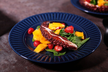 Octopus salad with mango and strawberries on a blue plate. Octopus tentacle on a blue plate with fruits and berries. Close-up.