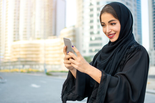 Portrait of beautiful smiling young Arabic woman in traditional abaya clothes using smartphone on the city street.