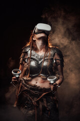 Fototapeta na wymiar Barbaric scandinavian woman fighter in light armour with virtual reality headset and joysticks poses in dark smokey background.
