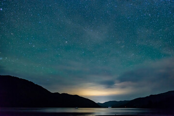 starry sky above the shape of mountains with the light of a city in a fjord in the Marlborough Sounds, New Zealand - obrazy, fototapety, plakaty