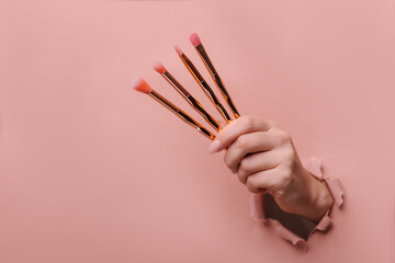 A female hand holds different makeup brushes. Hole in torn paper trendy beige background. Make-up...