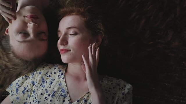 Haircare, skincare, beauty conception: two beautiful models, friends, sisters with long natural curly hair lay and touch their faces . Redhead and dark blonde curly models. Top view portrait.