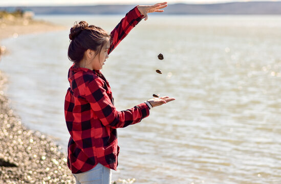 joven latina juega con piedras flotantes en la costanera con cielo azul y camisa a cuadros rojos y negros frente a un lago