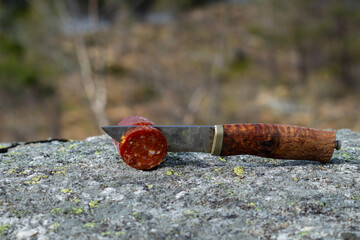 Knife cutting sausage in the mountain with blurry mountain landscape in background.  Outdoor food.