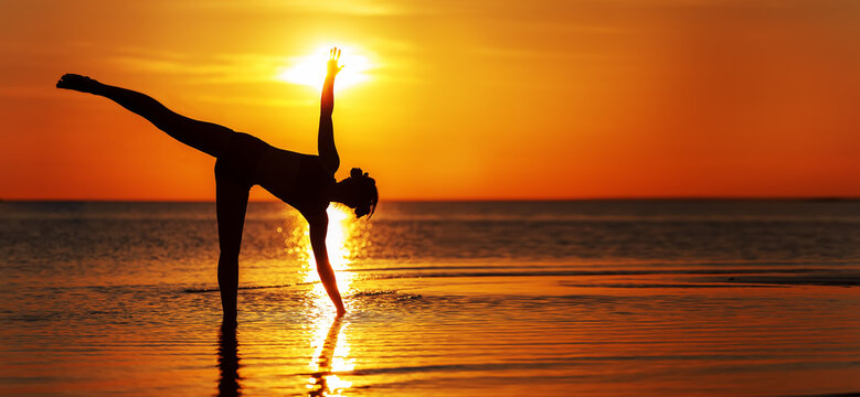 Fit sport girl demonstrates yoga asana Ardha Chandrasana (half moon pose) on the beach in the light of summer sunset. Wide image.