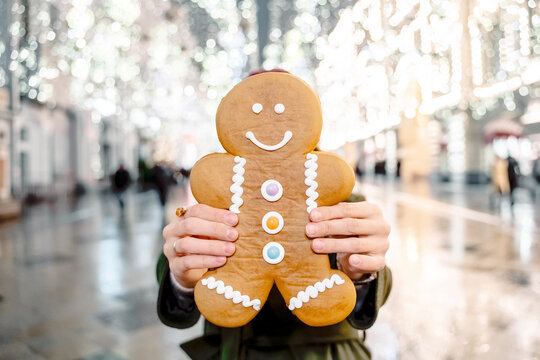 Young beautiful girl, woman with huge funny gingerbread man. Christmas shopping. Walking on market street in big city decorated with garlands, lights.Stylish look, wool coat, hat.New year celebration