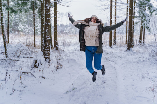 Young girl, woman walks is jumping in beautiful winter forest among trees, firs, covered with snow. Magnificent nature and views. Fashionable image, clothes, parka, hat, mittens, blue jeans