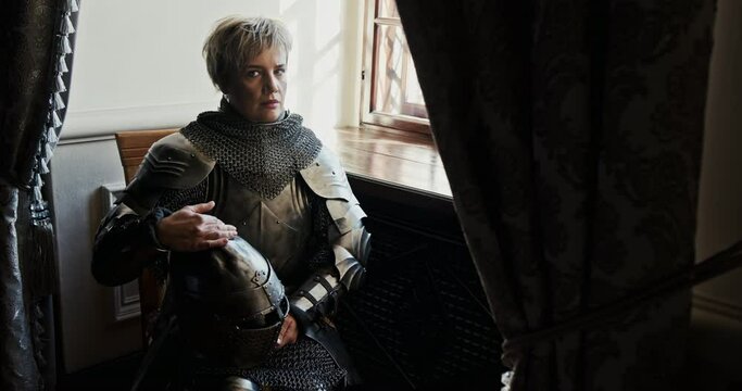 Adult woman in full armor looking out window and thinking about battle then turning head to camera while impersonating Joan of Arc. Medieval female warrior sitting near window