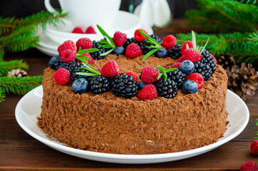 Chocolate honey cake with cream and fresh berries on top on a white plate on a wooden background. Cake for Christmas and New Year. Copy space.