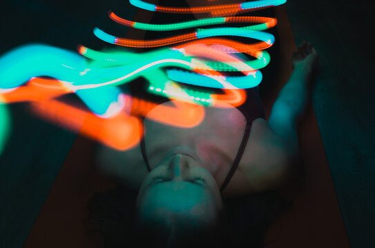 Young woman laying on yoga mat in shavasana with colourful light trails hovering above. Concept: wellness, energy healing, colour therapy, self care