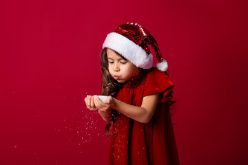 little girl in a Santa suit blows snow off her hands. red background isolate. space for text