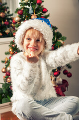 Beautiful little girl with santa claus hat sitting beside christmass tree. Thumb up