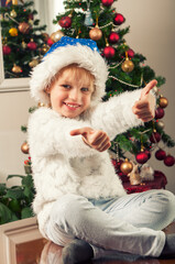 Beautiful little girl with santa claus hat sitting beside christmass tree. Thumb up