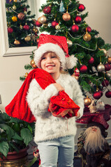 Beautiful little girl with santa claus hat and gift bag standing beside christmass tree