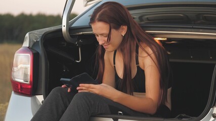 Young beautiful girl rides alone in the trunk of the car in the evening at sunset. Traveler in social networks phone. Work remotely on business trips. The modern woman is looking for adventure. Online