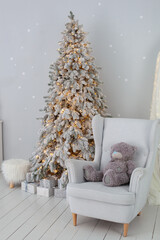 Cozy Scandinavian interior of room in light colors decorated with Christmas tree and armchair with plush teddy bear. New Year. Xmas gifts on the floor near Christmas snowy tree in living room. hygge	