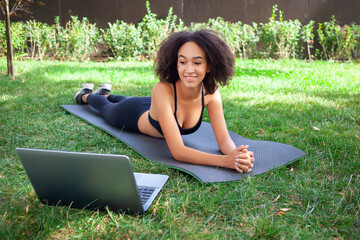 Cute fit dark-skinned girl lying on a sports mat in the backyard, smiling, looking at the laptop...
