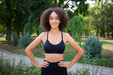 Attractive dark-skinned sportswoman holds hands on her belt, dressed in black sports top and leggings, smiling, stands against the green park background. People, sport and healthy lifestyle concept.