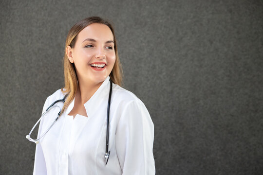 Horizontal close-up photo of attractive young female doctor in medical coat, looking far away and smiling broadly. Copy space isolated on gray background. Medicine, healthcare and people concept.